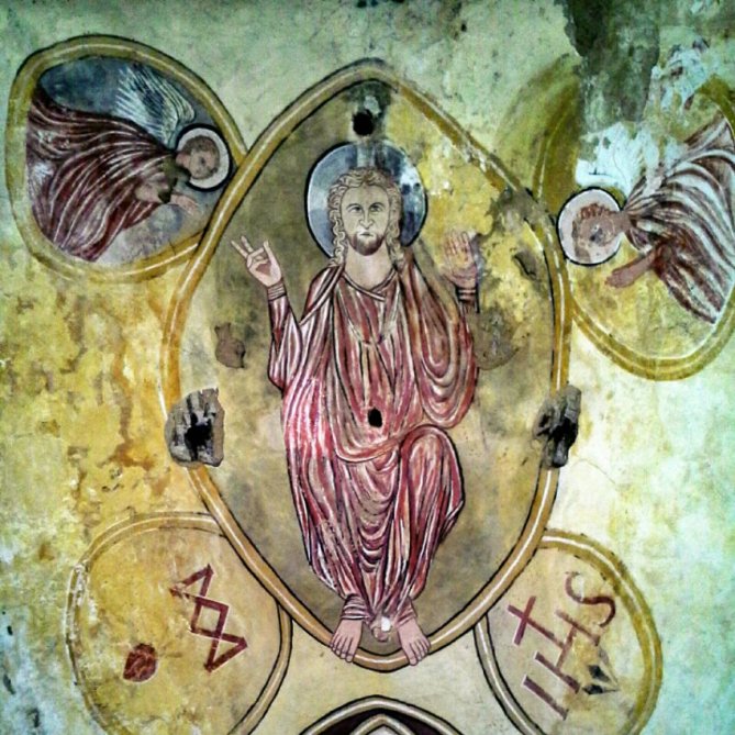 A faded mural depicting Christ seated in the center, with raised hands, flanked by two angels on either side, against a textured, weathered wall. Gargilesse Dampierre on the way to Santiago from Belgium