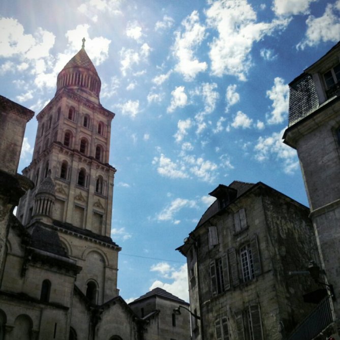 Uitzicht op een imposante kathedraal met een hoge toren, omgeven door historische gebouwen onder een heldere blauwe lucht met enkele wolken. Foto Jasmine Marie Josee Debels